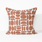 A square throw pillow featuring a modern geometric pattern of terracotta-colored abstract shapes on a white background, set against a clean white studio backdrop.