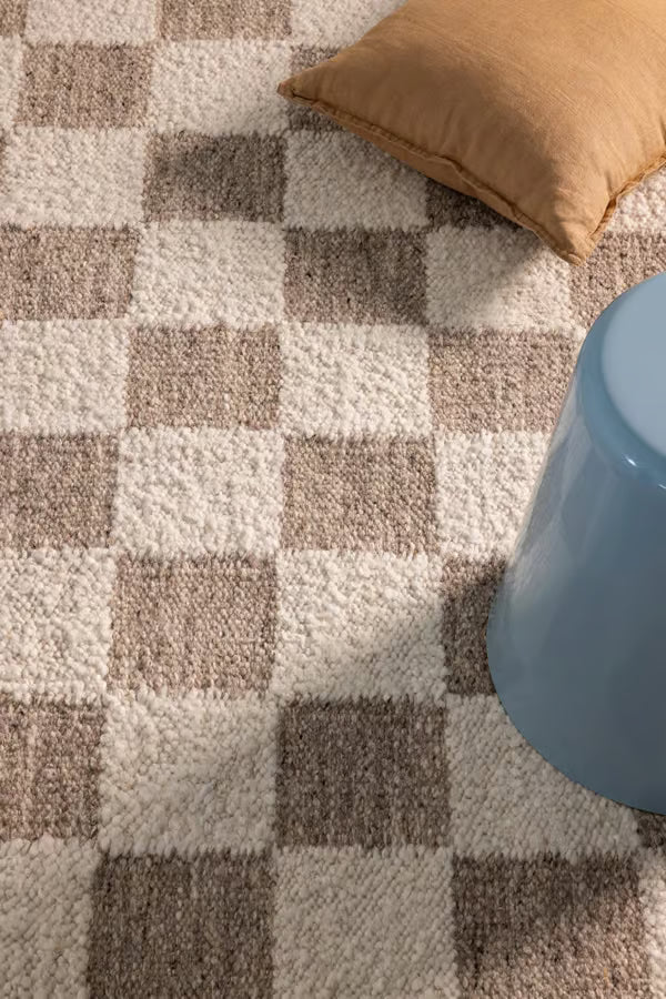 A close-up of a textured area rug with a beige, cream, and brown checkerboard pattern, styled with a tan pillow and a light blue stool.