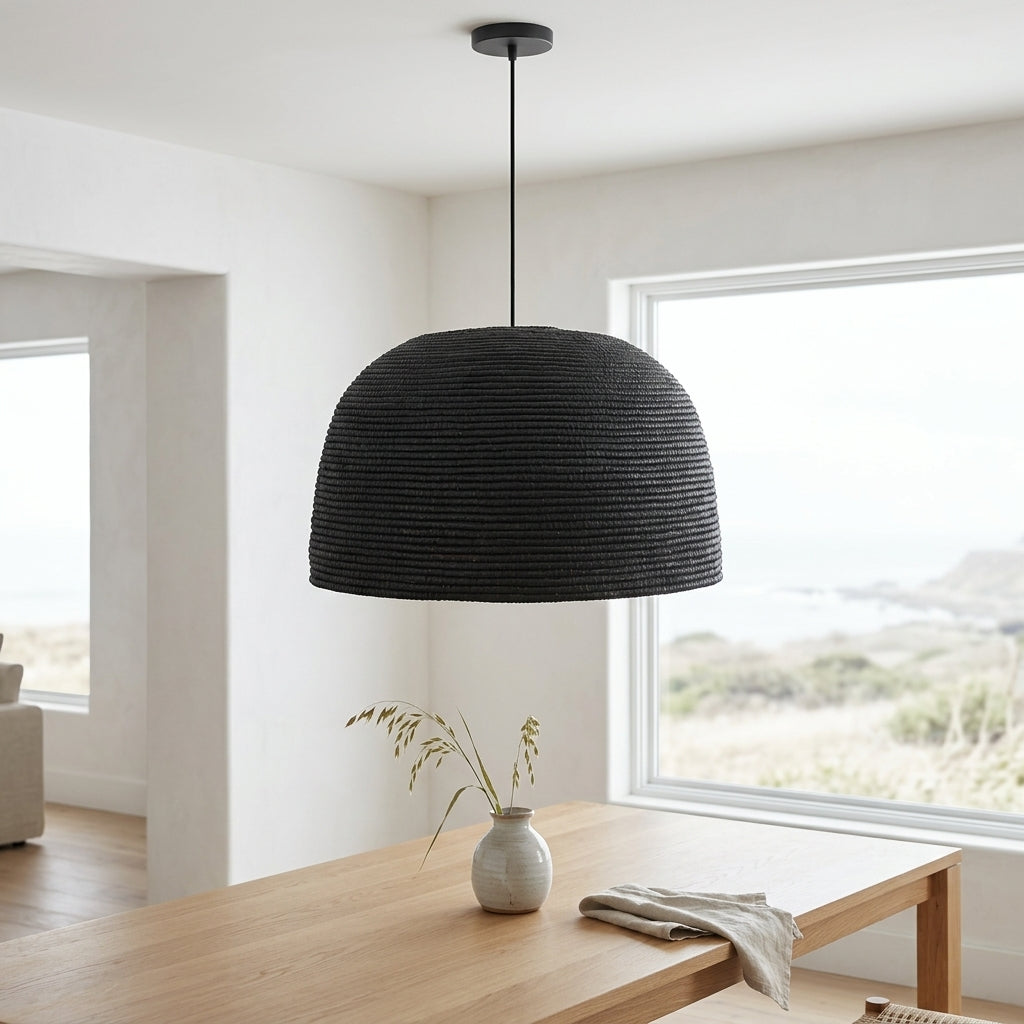 A black woven dome pendant light hanging over a light wood dining table in a bright, modern room with large windows.