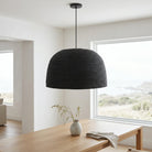A black woven dome pendant light hanging over a light wood dining table in a bright, modern room with large windows.