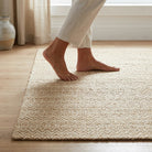 A person wearing light beige pants steps onto a textured cream and beige woven area rug on a light wood floor in a bright room.