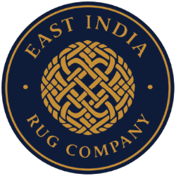 A circular navy blue and gold logo for East India Rug Company, featuring an intricate interwoven knot design in the center with the company name arched above and below.