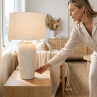 A woman in a beige linen pajama set reaches toward a textured cream ceramic table lamp on a light wood nightstand in a bright, modern bedroom.