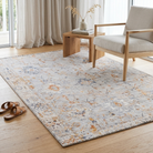 A rectangular area rug with a distressed, vintage-style floral pattern in muted blue, orange, and beige tones, styled on a light wood floor in a bright living room next to a wooden armchair and side table.