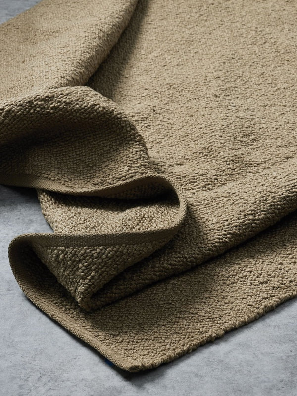 A close-up of a folded, textured woven fabric in a warm taupe-brown color, showing its chunky knit pattern against a neutral gray background.