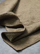 A close-up of a folded, textured woven fabric in a warm taupe-brown color, showing its chunky knit pattern against a neutral gray background.