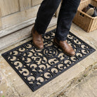 Person wearing brown shoes stepping onto a decorative black doormat with a wooden door and wicker basket in the background.