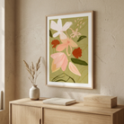 A framed botanical art print featuring pink, white, and rust-red flowers on an olive green background, hanging on a textured beige wall above a light wood console table with a vase and books.