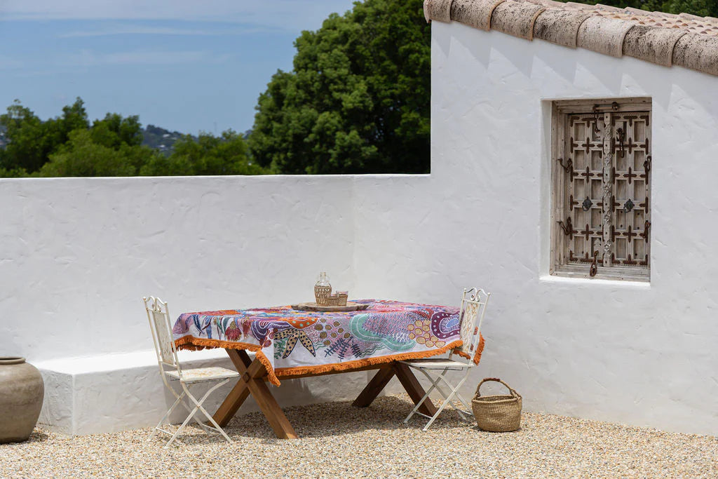 Outdoor setting with a table and chairs under a colorful tablecloth, against a white wall with a window.