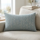 A rectangular throw pillow in a textured slate blue fabric with subtle white flecks, resting on a light beige linen sofa in a bright, modern living room.