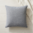 A square gray textured throw pillow resting on a light wood floor next to a cream linen fabric.