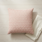 A square blush pink textured throw pillow resting on a light wood plank floor next to a folded cream linen napkin.