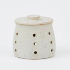 A small ceramic diffuser or candle holder with a lid, featuring a speckled off-white glaze and multiple circular ventilation holes on the side, set against a plain white background.
