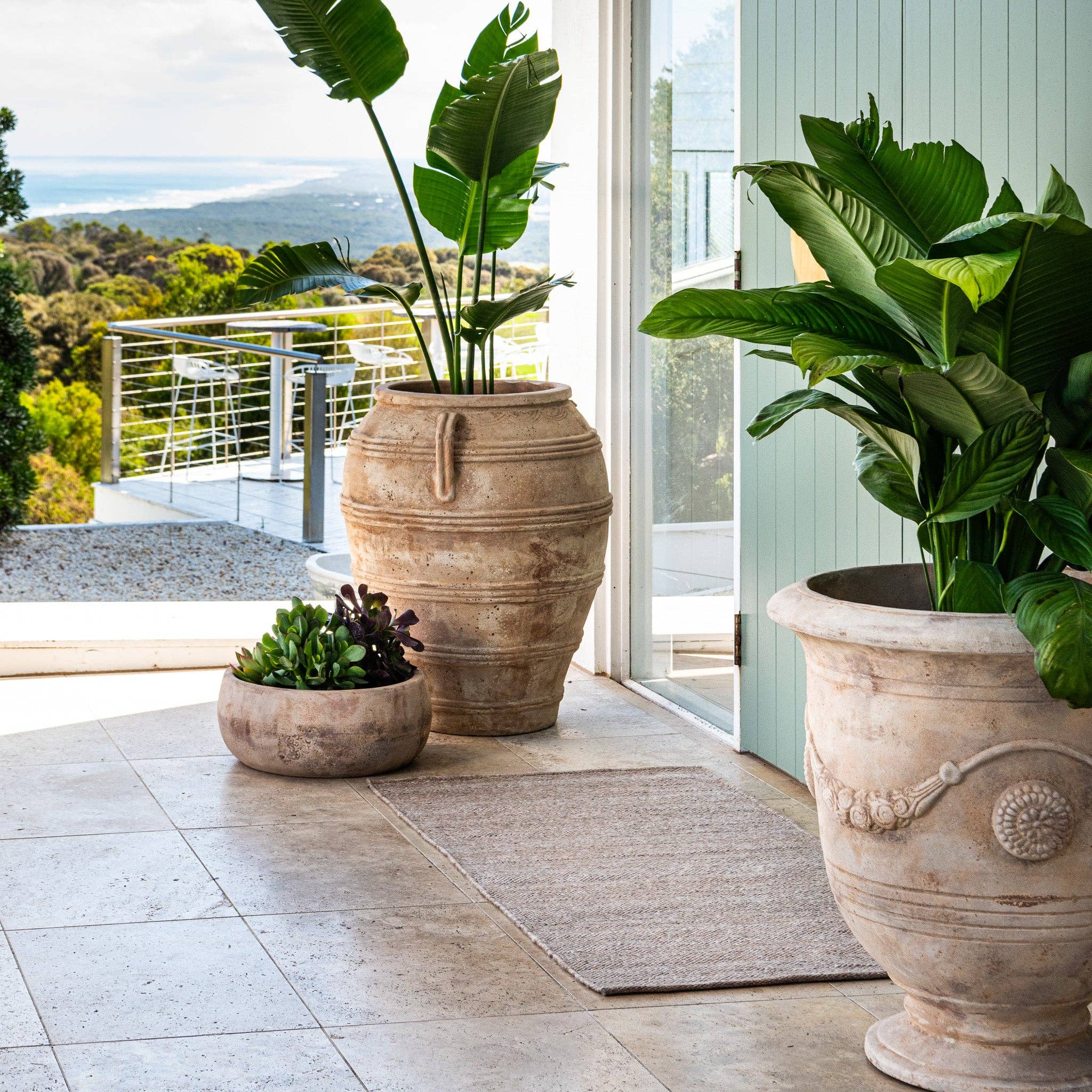Three large decorative pots with plants on a patio with a scenic view.