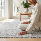 Person sitting on a textured rug in a bright room with large windows and a wooden table.