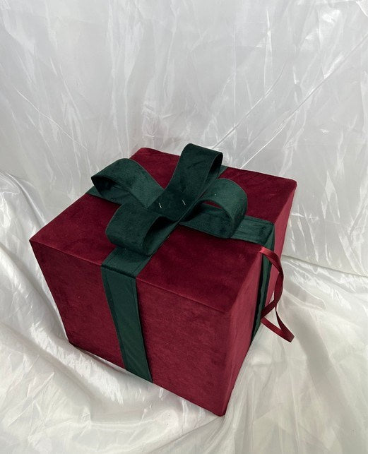 A square gift box wrapped in deep red velvet fabric and tied with a dark green satin ribbon and bow, set against a wrinkled white backdrop.