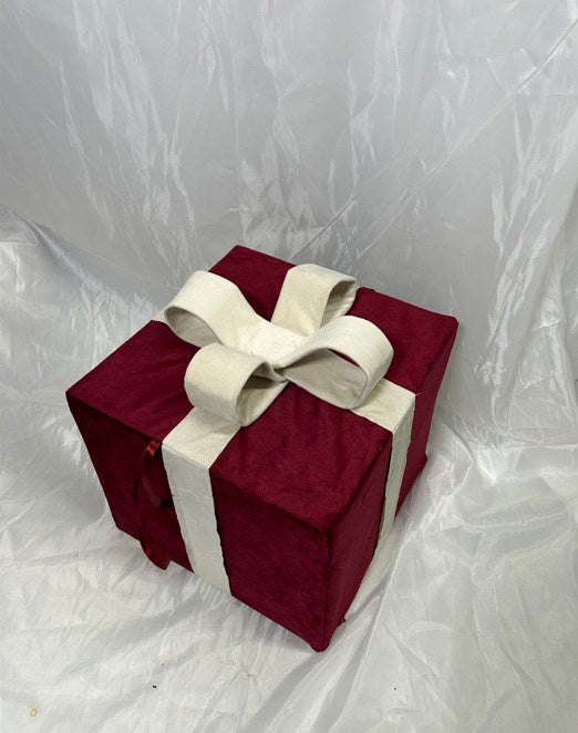 A square gift box wrapped in deep burgundy velvet fabric with a wide cream ribbon tied into a large bow on top, photographed against a wrinkled white backdrop.