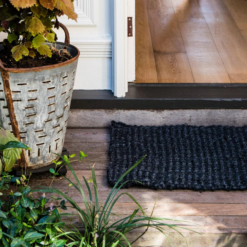 Black doormat on a wooden floor with plants and a metal basket in the foreground.