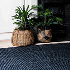 Two potted plants in woven baskets on a blue textured rug in front of a fireplace.