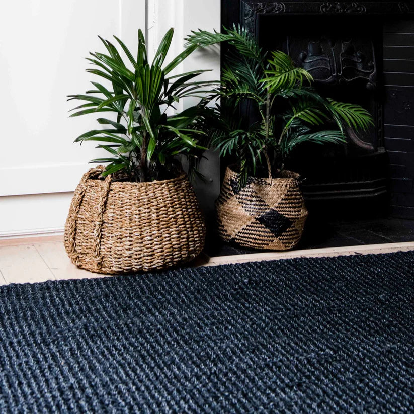 Two potted plants in woven baskets on a blue textured rug in front of a fireplace.
