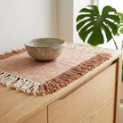 Ceramic bowl on a woven placemat with fringe on a wooden surface, with a plant in the background.