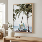 A framed vertical beach art print featuring palm trees and three colorful surfboards on a wooden console table in a bright, modern living room.