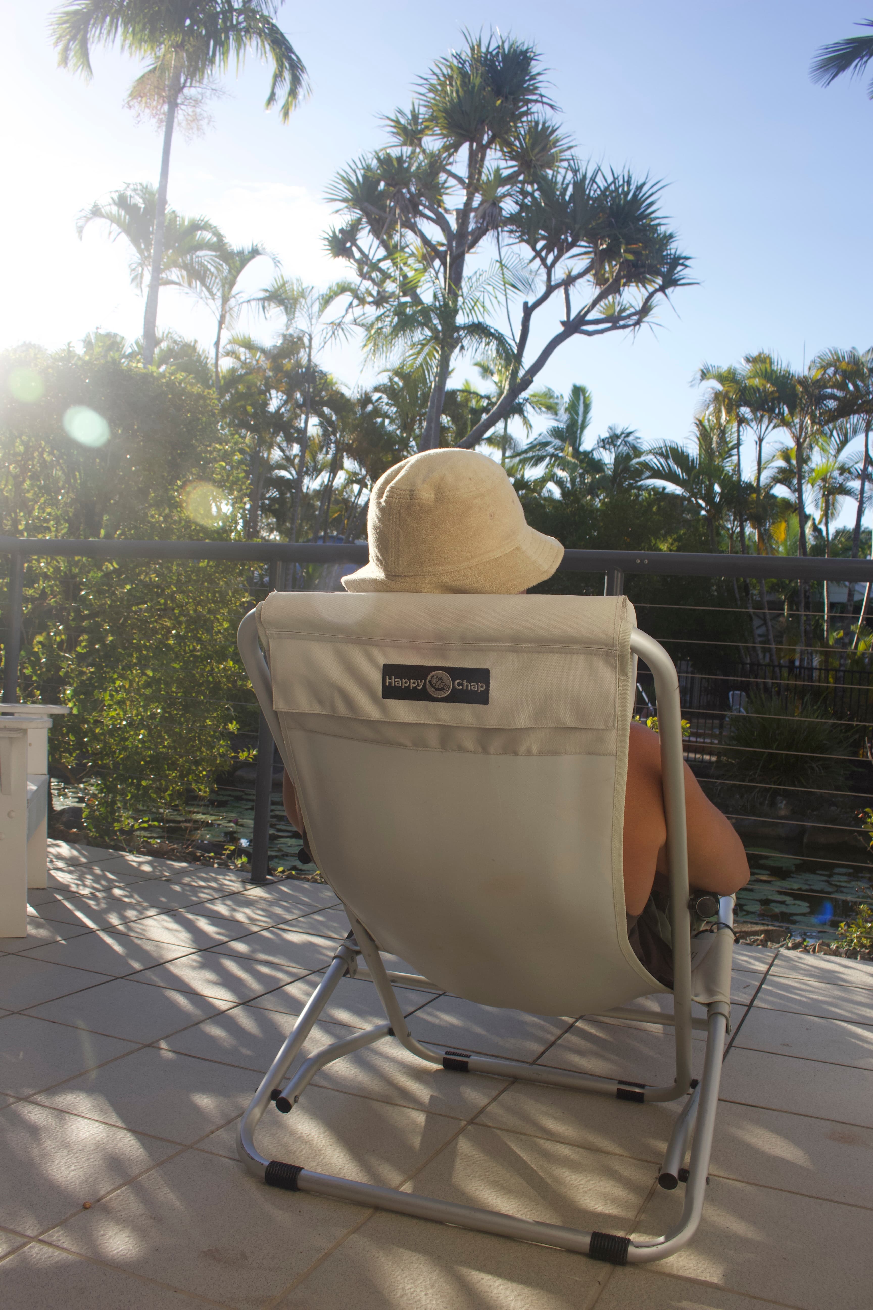 Person sitting in a beige chair with a logo, wearing a wide-brimmed hat, on a tiled patio with palm trees in the background.