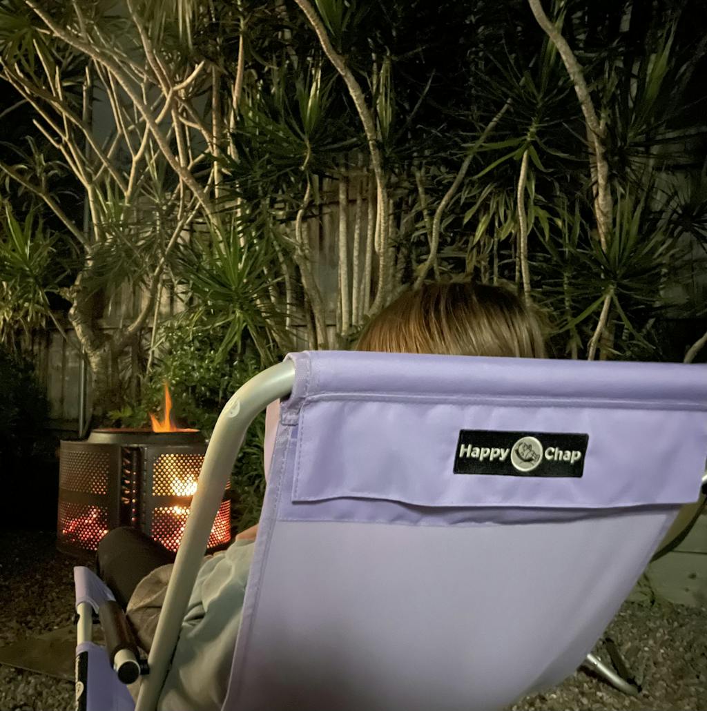 Person sitting in a purple chair with 'Happy Chap' logo near a fire pit at night.