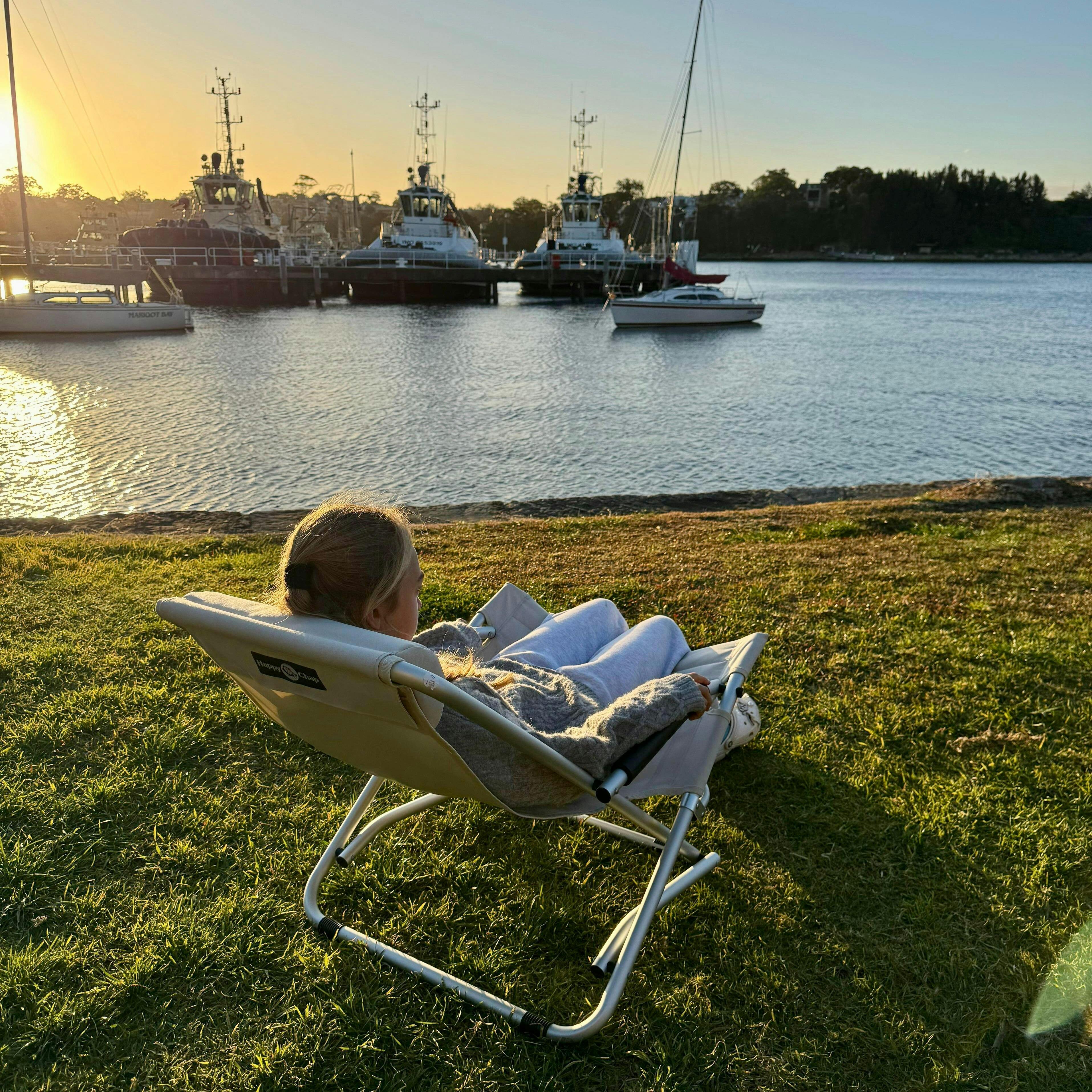 Person sitting in a chair by a waterfront with boats in the background at sunset.
