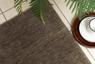A rectangular emerald grey wool rug placed on a tiled floor with a round wooden object and green foliage in the background.