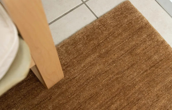 Close-up view of a honey brown wool rug with a textured, chunky woven style, placed on a tiled floor next to a wooden furniture leg.