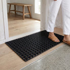Person stepping onto a black doormat with a wooden bench in the background.