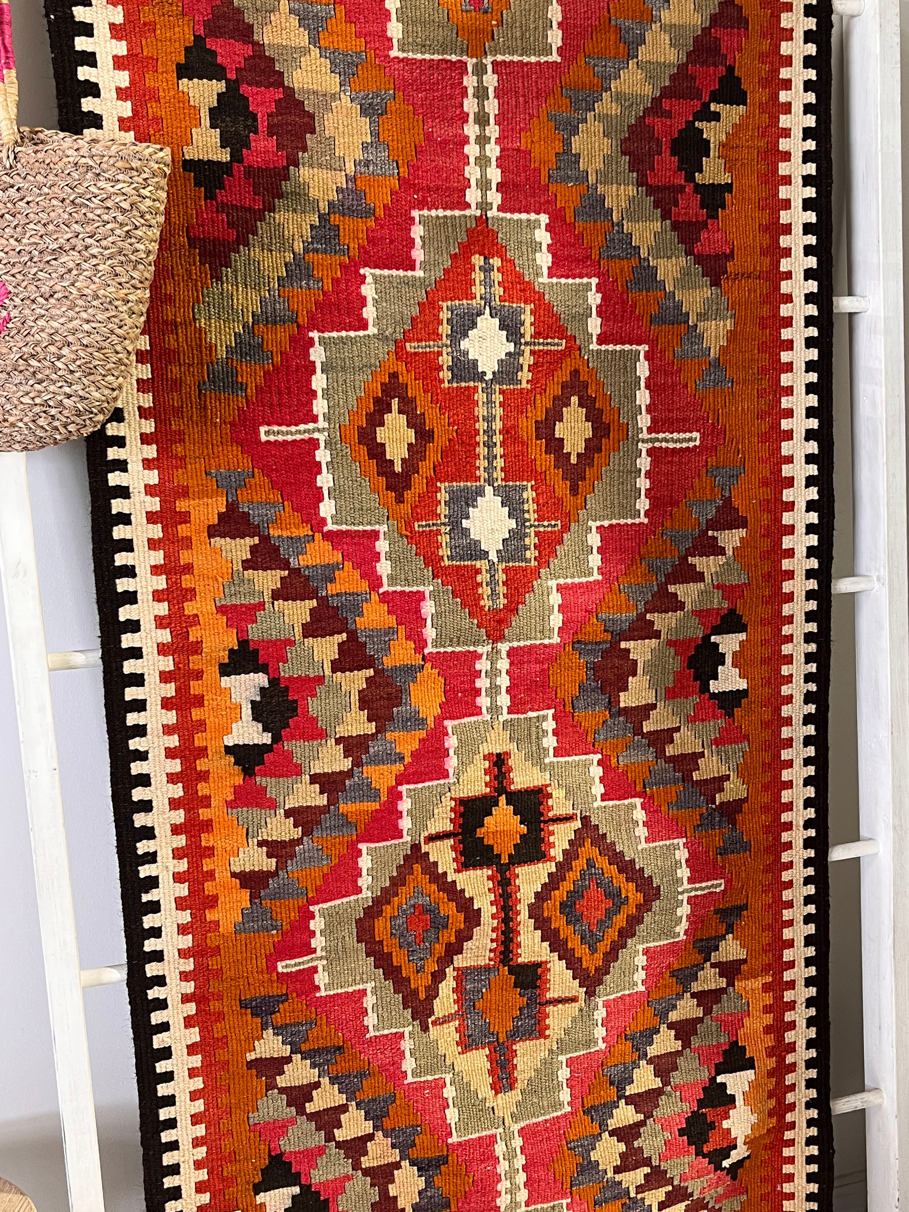 A close-up of a woven geometric runner rug with a Southwestern or Navajo-inspired pattern in red, orange, beige, brown, black, and gray, draped over a white ladder with a woven straw bag hanging on the left.