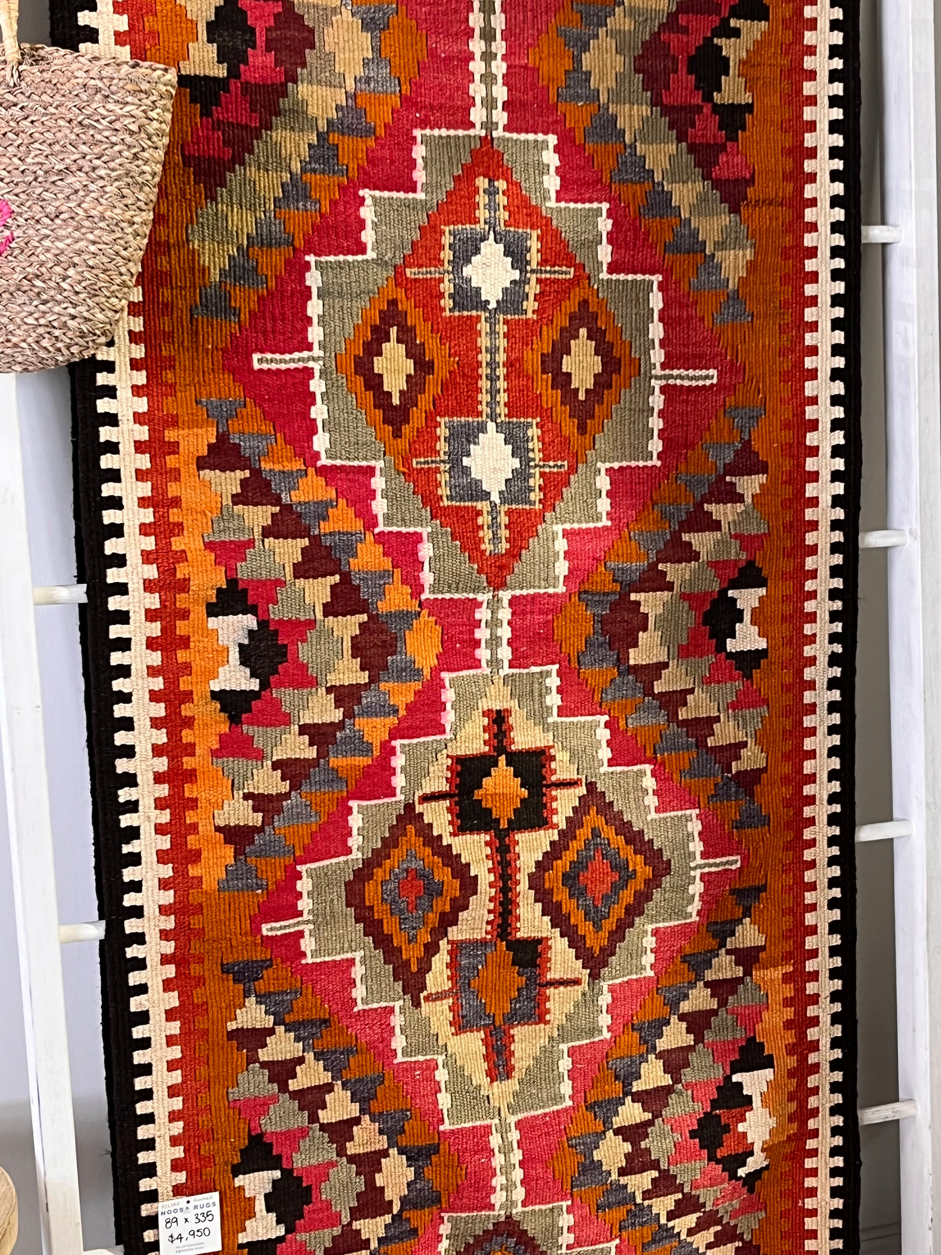 A vintage-style kilim runner rug with a geometric tribal pattern in red, orange, brown, beige, and black, hanging on a white rack.