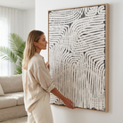 A woman in a beige linen outfit stands in a bright living room, holding a large framed abstract canvas print with black curved lines on a white background.