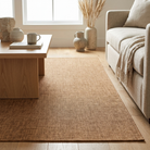 A natural jute area rug in a warm tan-brown color, styled in a bright, modern living room next to a light beige sofa and a wooden coffee table.
