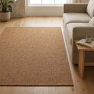 A rectangular natural jute area rug in a warm tan-brown color, shown in a bright, modern living room setting next to a light beige sofa and a small wooden side table.