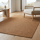 A rectangular natural jute area rug in a warm tan-brown color, styled in a bright, modern room with light wood floors, a beige armchair, and a wooden side table.