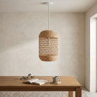 A natural woven rattan pendant light hangs over a wooden table in a minimalist, warm-toned room with a textured beige wall.
