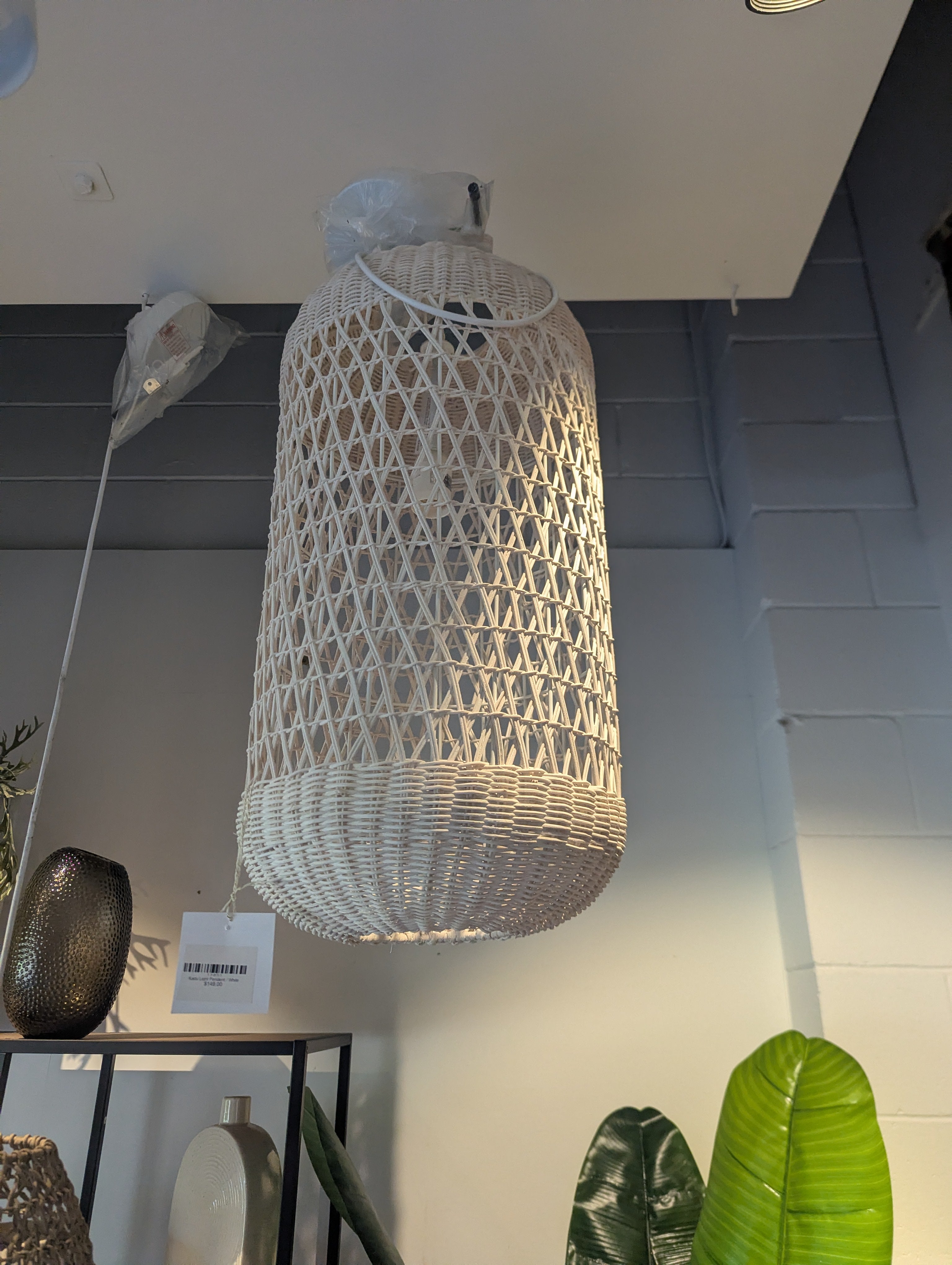 A large cylindrical pendant light made of woven natural fiber, hanging from a ceiling in an indoor showroom. The lamp has an open lattice top and a tightly woven lower shade.