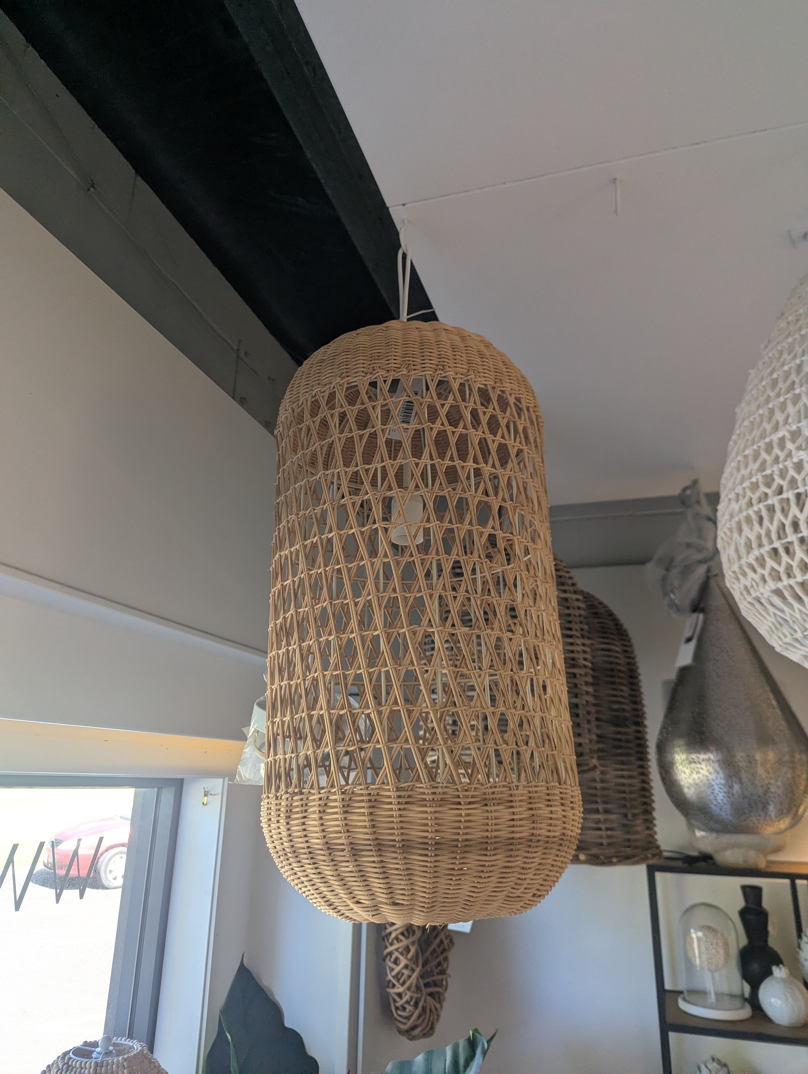 A large cylindrical woven rattan pendant light hanging in a modern room with a white ceiling and dark wooden beams.
