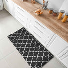 A black rectangular kitchen mat with a white geometric trellis pattern lies on the floor in front of white cabinets with light wood countertops. The scene is styled like a modern kitchen with jars of pasta and bread on the counter.