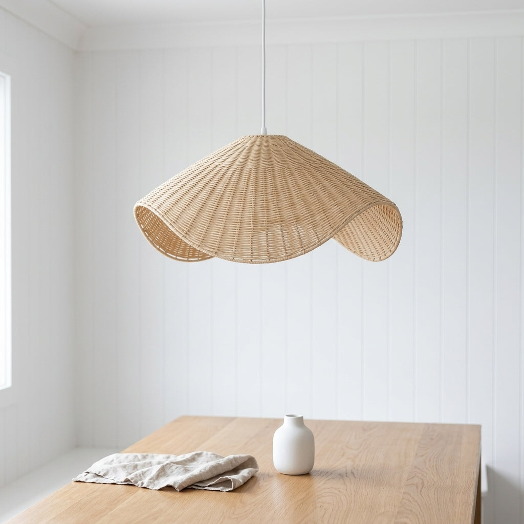 A natural woven rattan pendant light with a scalloped, wavy silhouette hangs over a light wood table in a bright, white-paneled room. A small white ceramic vase and a folded linen cloth sit on the table.