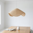 A natural woven rattan pendant light with a scalloped, wavy silhouette hangs over a light wood table in a bright, white-paneled room. A small white ceramic vase and a folded linen cloth sit on the table.