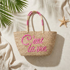 A woven straw tote bag with pink embroidered text reading 'C'est la vie' and pink-and-cream handles, styled on a light fabric surface with palm leaves and a starfish.
