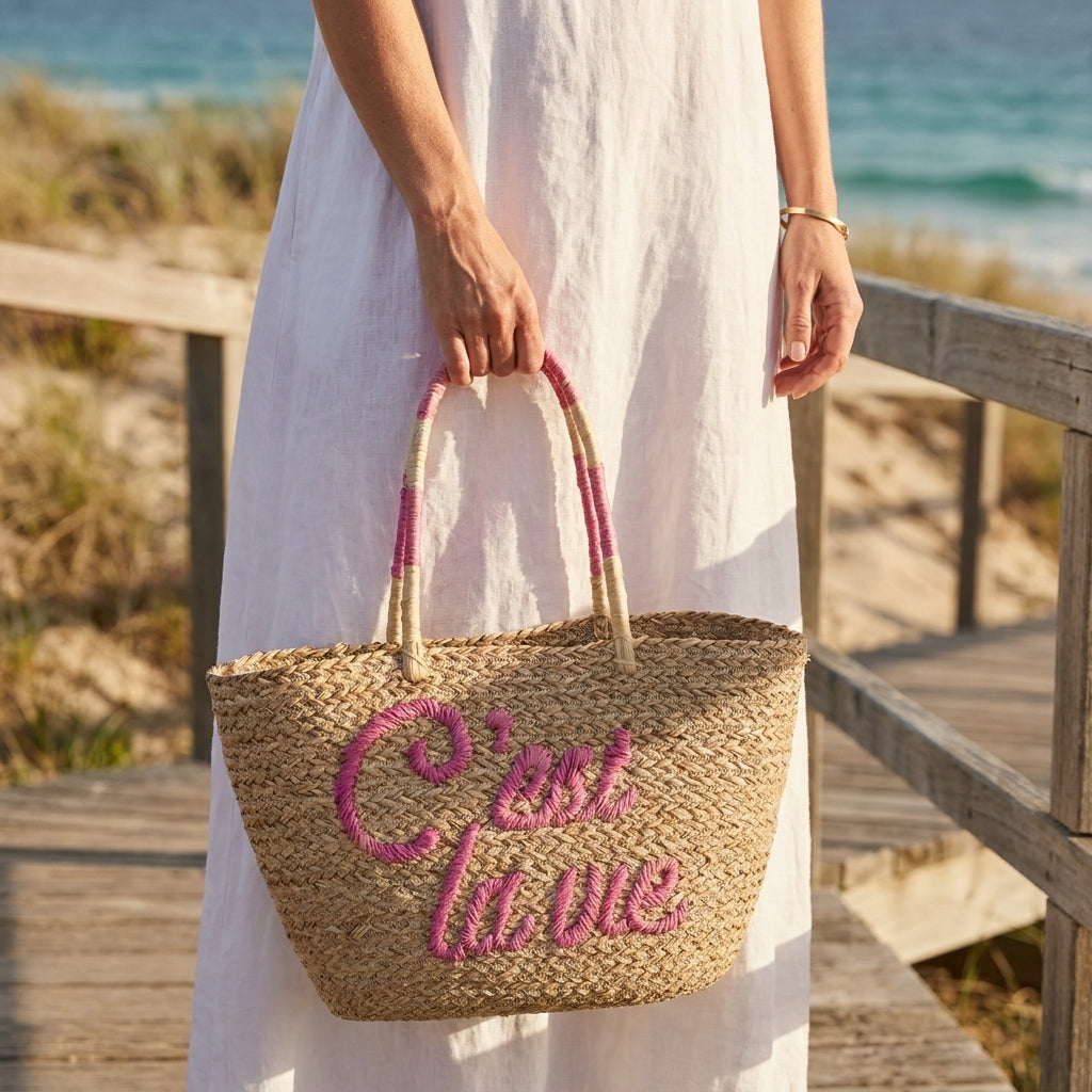 A woman in a white dress holds a woven straw tote bag with pink embroidered text reading "C'est la vie" and pink-and-cream handles, photographed outdoors on a wooden boardwalk near the beach.