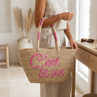 A woman in a light linen dress holds a natural woven straw tote bag with pink embroidered cursive text reading 'C'est la vie' and pink-and-beige striped handles, standing in a bright, neutral-toned room next to a wooden table.