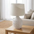 A white woven rattan table lamp with a beige linen drum shade sits on a light wood side table in a bright, modern living room setting.