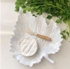 White leaf-shaped dish with a round item and knife, surrounded by green leaves on a light background