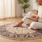 A woman sits barefoot on a round, vintage-style multicolor area rug in a bright, modern room, holding an open book. The room features light wood floors, beige curtains, and a potted plant in the background.